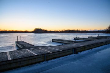 Fototapeta premium wide angle shot of wooden boat docks on icy blue frozen lake with warm orange and yellow sunset on the horizon