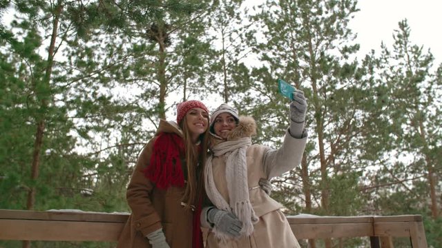Two young pretty women smiling at camera while standing outdoors on wooden terrace in pine forest, taking selfie with smart phone at winter day and then watching photos