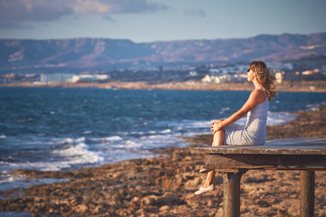 Young woman with long hair sitting at the edge of wooden pier and watching sunset at the rocky coast of Paphos, Cyprus