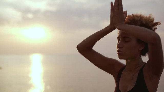 Focuses young dark-skinned female doing meditation with folded hands near sea. Silhouette