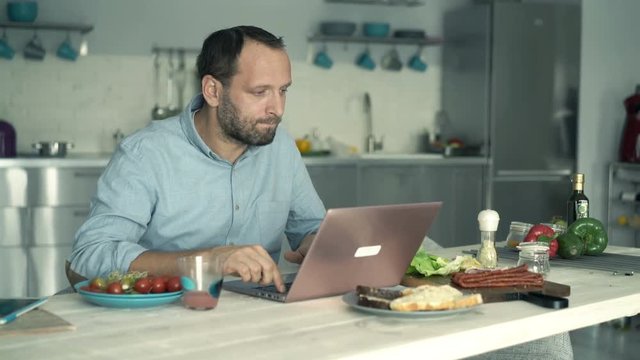 Man with laptop having head pain during breakfast in kitchen at home
