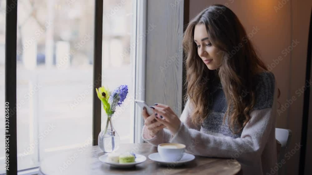 Wall mural Beautiful young woman sitting at cafe and waiting for her boyfriend while texting on mobile phone