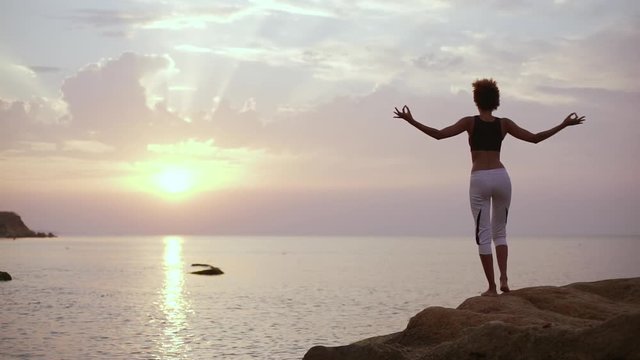Dark-skinned female with afro hairstyle standing facing sun and sea doing yoga in slowmotion. Silhouette