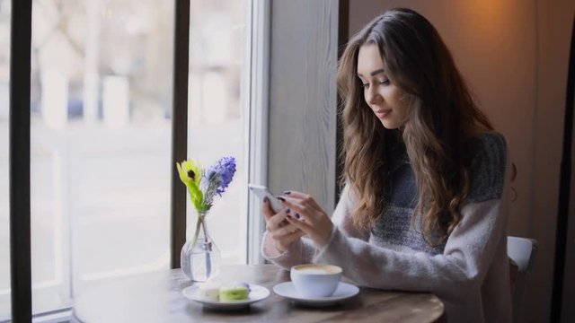 Beautiful young woman sitting at cafe and waiting for her boyfriend while texting on mobile phone