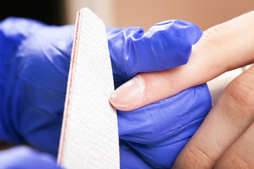 Manicure specialist care by finger nail in beauty salon.  Finger closeup isolated on blue gloves background. Manicurist uses professional manicure tool.