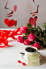 Closeup of cup of coffee with the bouquet of red roses of Valentine's day. Yellow sticky note with the date February 14.