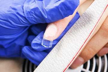 Fototapeta na wymiar Manicure specialist care by finger nail in beauty salon. Manicure in beauty shop. Gel nails. Finger closeup isolated on blue gloves background. Manicurist uses professional manicure tool.