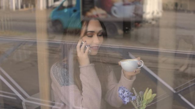 Smiling beautiful young woman drinking coffee and talking on mobile phone at the window in cafe