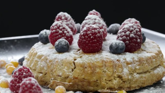 Cake with raspberries, blueberries, sea buckthorn sprinkled with powdered sugar on a black plate rotates