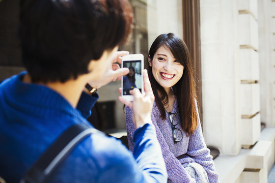 Young Japanese Man And Woman Enjoying A Day Out In London, Standing On A Pavement, Taking A Picture.