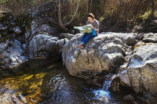 A woman sitting on the rocks in the shade by a river, looking at a map. 