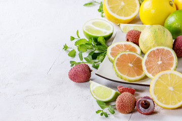 Fototapeta na wymiar Variety of whole and sliced citrus fruits pink tiger lemon, lemon, lime, mint and exotic lichee on white square plate over white concrete textured background. Copy space. Healthy eating