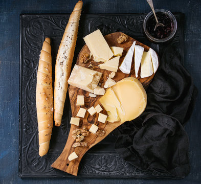 Cheese plate. Assortment of cheese with walnuts, jam and bread on olive wood serving board with textile over black ornate and dark blue canvas as background. Top view with space.