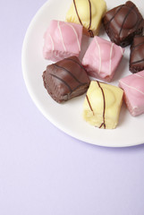 A plate full of fondant fancies cakes on a pastel purple background with blank space below
