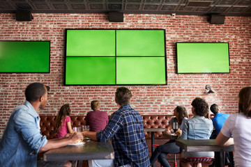Rear View Of Friends Watching Game In Sports Bar On Screens