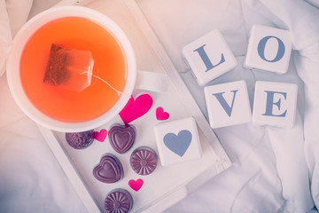 Breakfast in bed in Valentines day. Cup of tea and chocolate sweet candies. Message love spelled in wooden blocks and paper hearts. Top view with copy space