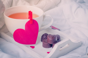 Breakfast in bed in Valentines day. Cup of tea and chocolate sweet candies. Message on paper hearts. Copy space