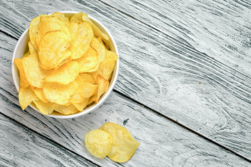 Potato chips in bowl on a wooden background, top view. Salty crisps scattered on a table.
