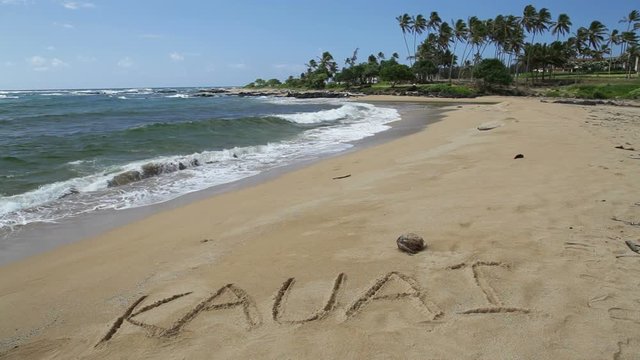 The word Kauai written in the sand on a beach on the island of Kauai, Hawaii. A coconut is visible above the letter A. 