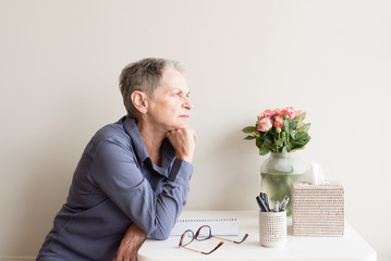 Older woman with short grey hair and blue shirt seated at desk with hands on chin looking pensive (selective focus)