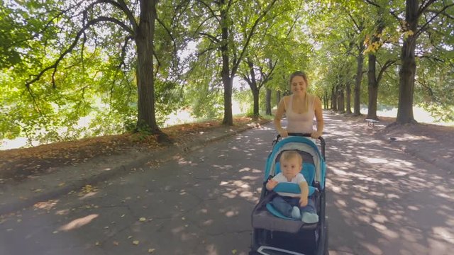 Steadicam shot of happy smiling mother walking with baby pram on alley at park