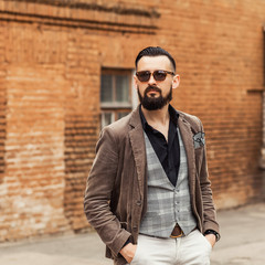 Young confident hipster man with beard in glasses posing on the street in old town. Cute man wearing a vest, black shirt and light trousers. Man keeps hands in pockets and looking forward