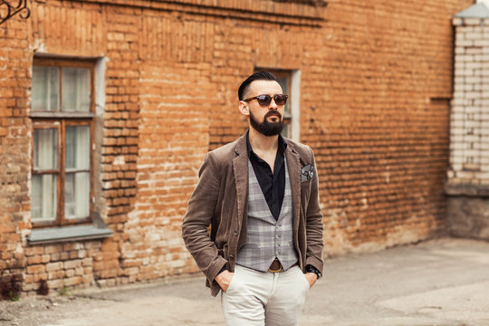 Young confident hipster man with beard in glasses posing on the street in old town. Cute man wearing a vest, black shirt and light trousers. Man keeps hands in pockets and looking forward