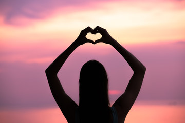 Woman holding up shape of a heart against a beautiful sunset. 