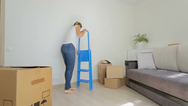 Beautiful young woman putting stepladder next to empty wall at new apartment