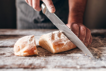 Cutting fresh crusty bread close-up. Baker carving loaf of warm soft tack with big knife on kitchen...