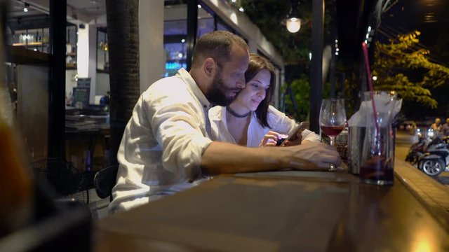 Young couple talking and browsing web on smartphone sitting in cafe at night
