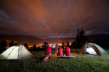 Friends sitting on a bench made of logs and watching fire together beside camping and tents in the night on the background mountains and luminous town. Rear view