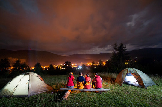 Friends sitting on a bench made of logs and watching fire together beside camp and tents in night on the background mountains and luminous town. Rear view
