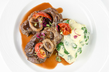 Braised beef on the bone with dried tomatoes and mashed potatoes with spinach and feta cheese on a plate on a white background, top view