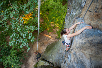 Brave woman rock climber is climbing with rope on a rocky wall. Belayer standing on the ground insuring the climber. View from the top. Extreme sports concept