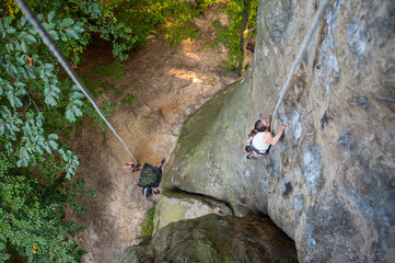 Young woman rock climber is climbing with rope on a rocky wall. Belayer standing on the ground insuring the climber. View from the top