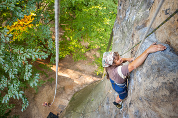 Older athletic woman rock climber is climbing with carbines and rope on a rocky wall. Belayer standing on the ground insuring the climber. View from the top. Extreme sports concept