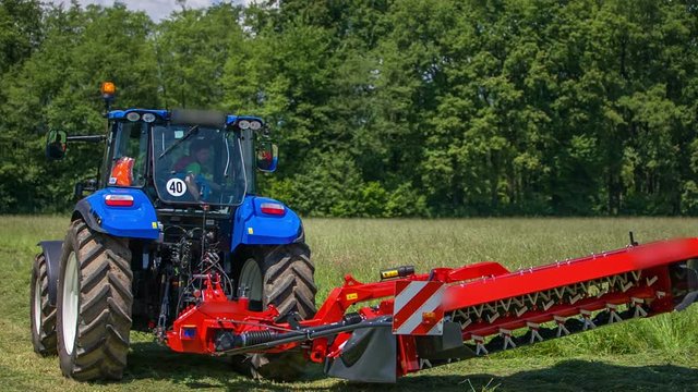 A farmer in a blue tractor pushes a button and machinery for cutting grass lowers down to the ground.
