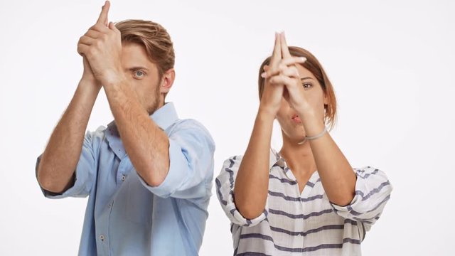 Cheerful Caucasian pair standing back to back playing imagine guns and pointing them to camera on white background
