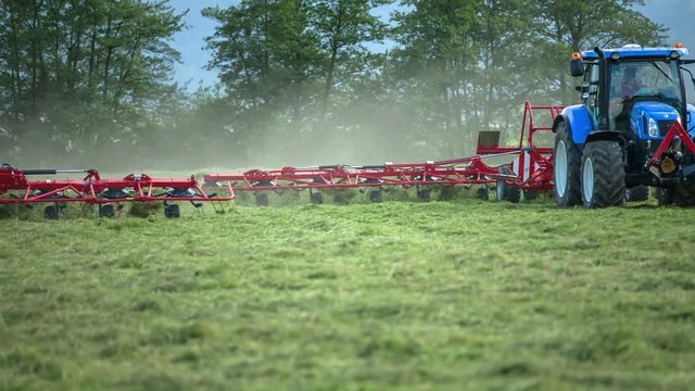Three tractors are working outside on the grass-fields and are organizing freshly cut grass with rotary rakes.
