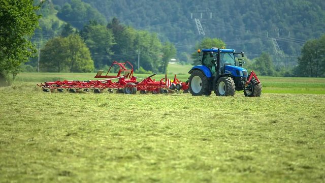 A blue tractor is preparing hay. With the help of the agricultural machinery it is turning hay around.
