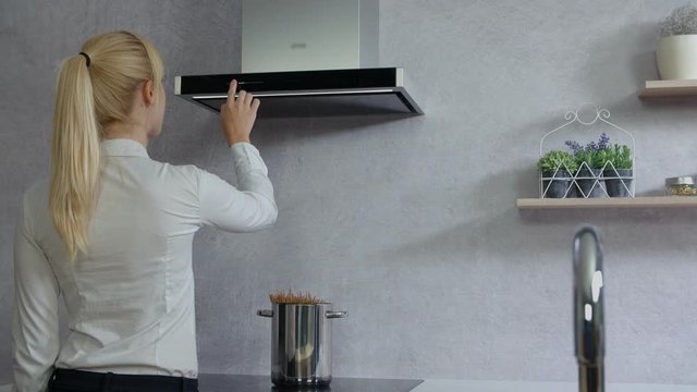 We can see a pot with spaghetti being cooked on the stove. A young woman comes into the kitchen and she adjust a few buttons on the kitchen hood.
