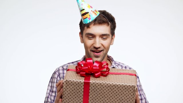 Happy young Caucasian male with dark hair and light bristle wearing festive cap recieving birthday present box on white background