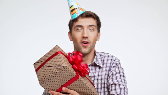 Happy young Caucasian male with dark hair and light bristle wearing festive cap recieving birthday present box on white background in slowmotion