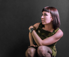 Sexy military girl in camouflage dress sits in crouched position and looks sideways