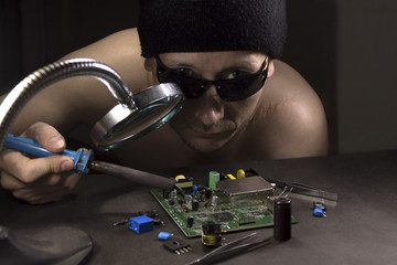 A man in a black hat and sunglasses with a soldering iron mounts radio-electronic circuits at the table.