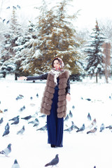 милая и красивая девушка брюнетка, кормит птиц в парке, вокруг лежит снег, зима, одета в длинное платье, шубу и платок с узорами