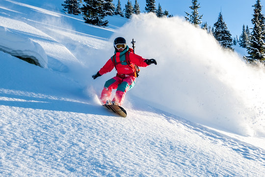 Young lady snowboarder in pink overall riding down on splitboard splashing snow