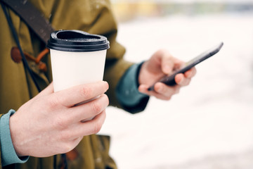 Man holding a cup of coffee in their hands and looking at phone on the street in winter