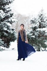 красивая, молодая девушка брюнетка гуляет в парке возле снежных елок, одета в длинное шифоновое платье и шубу